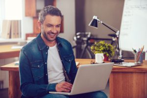 Confident young man working on laptop and smiling while sitting at his working place in office